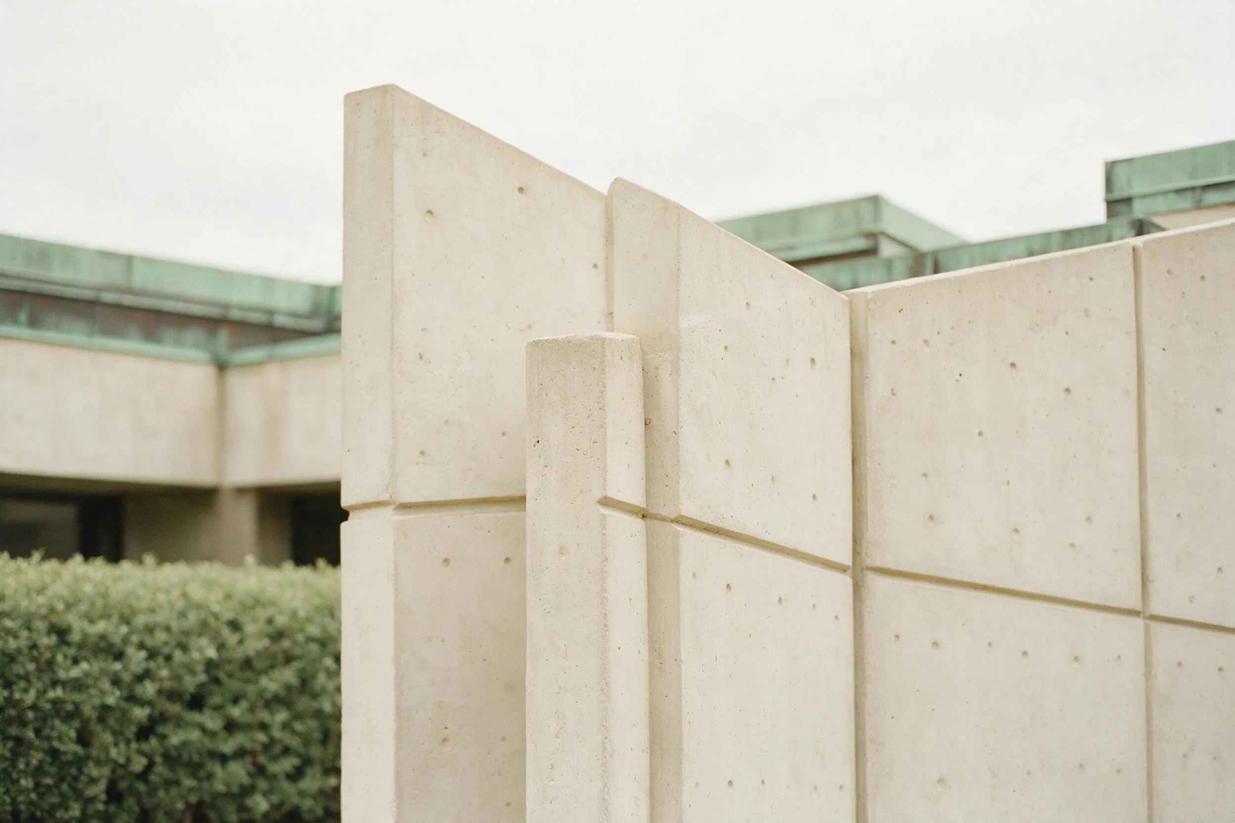 Precast concrete facade panels meeting at angles, clipped hedge and weathered copper flashing behind